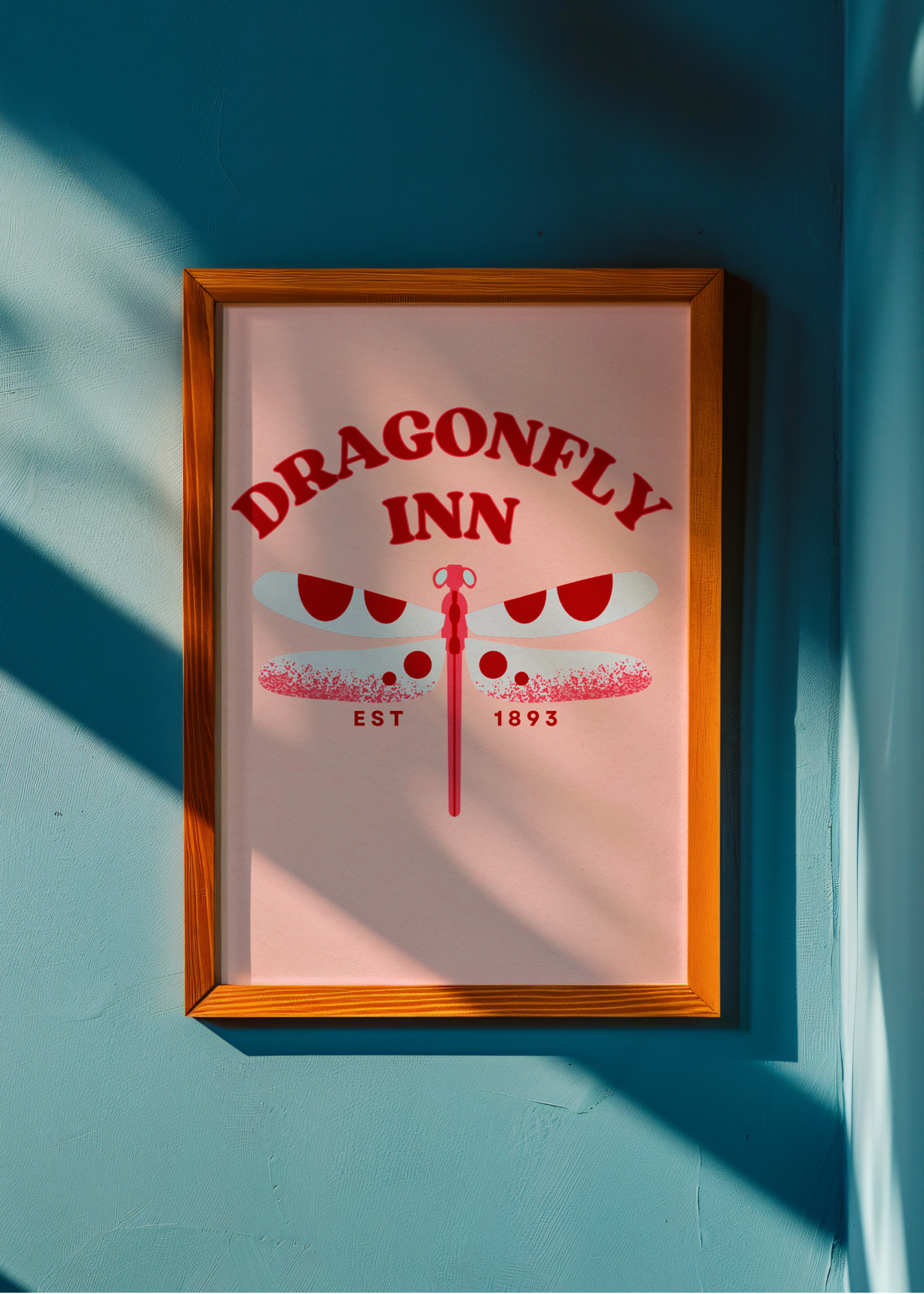 Framed poster of the Dragonfly Inn with a red dragonfly design on a light pink background, against a blue wall.
