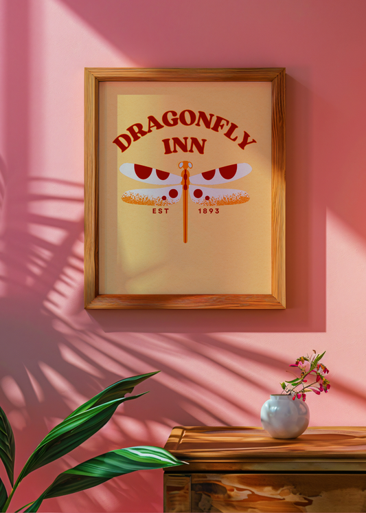 Framed print of 'Dragonfly Inn' on a pink wall with a vase and plant below.